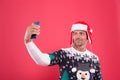 Merry christmas. cheerful unshaven man in santa hat and sweater making selfie on phone. man celebrate xmas party. winter