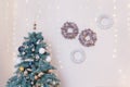 Merry Christmas fir-tree decorated with white and golden baubles, wreaths on white wall with shiny garland lights bokeh. Royalty Free Stock Photo