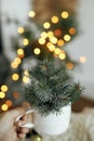 Merry Christmas! Hand holding stylish cup with fir branches on background of cozy knitted sweater, warm lights and sweden star in