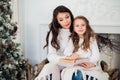 Merry Christmas and Happy Holidays, Pretty young mom reading a book to her cute daughter near tree indoors. Royalty Free Stock Photo