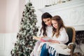 Merry Christmas and Happy Holidays, Pretty young mom reading a book to her cute daughter near tree indoors. Royalty Free Stock Photo