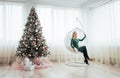 Merry Christmas and Happy New Year. Beautiful blonde woman in a green dress sitting in chair suspended at the tree Royalty Free Stock Photo