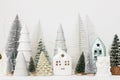 Merry Christmas! Stylish little Christmas trees and house decorations on white table. Modern christmas scene, miniature snowy