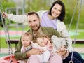 Merry family with two children having fun on outdoor playground. Young parents rides daughters on swing. Spring/summer/autumn Royalty Free Stock Photo