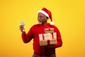 Merry senior black man in Santa hat holding Christmas gifts and credit card, doing Xmas shopping on orange background