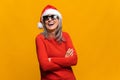 Merry senior middle-aged woman wearing santa hat stands with arms crossed