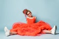 Merry smiling girl with colorful dreadlocks in coral big dress, sneakers, sunglasses sits on floor legs wide apart Royalty Free Stock Photo