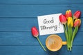 Message GOOD MORNING, tulips and coffee on blue wooden table, flat lay. Space for text