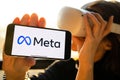 Meta logo on the iPhone, rebranding. Woman holding smartphone with new facebook logo wearing Oculus VR headset.