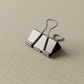 A metal binder clip sits on a textured beige surface. The clip is chrome-colored, with tw Royalty Free Stock Photo