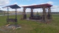 A metal brazier with a canopy and a wooden gazebo with benches, tables and a plastic roof stand on a planked area surrounded by la