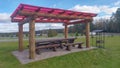 A metal brazier with a canopy and a wooden gazebo with benches, tables and a plastic roof stand on a tiled area surrounded by lawn