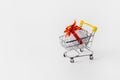 Metal cart from a supermarket with a gift on a white background. The gift is wrapped in white paper, tied with a red ribbon. The Royalty Free Stock Photo