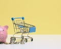 Metal miniature shopping cart with change and pink piggy bank on a white table, yellow background. Concept of saving budget, Royalty Free Stock Photo