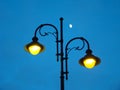 A metal pole is installed on a city street. On it are two electric street lights. In the evening twilight, half of the moon is alr Royalty Free Stock Photo