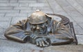 A metal statue of a plumber peering out of an open sewer hatch and looking at passers-by from the road level. Bratislava