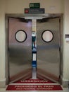 Metal swing door with two round windows in hospital and signs that say in Spanish: emergency exit, urgencies, prohibited passage,