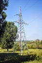 Metallic pylon of high-voltage power lines against blue sky. Located among trees with green foliage. Royalty Free Stock Photo