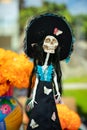Mexican skeleton figure in national headdress and blue and black dress isolated on the background of a bouquet of orange flowers.