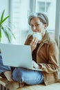 Mid aged businesswoman takes off her mask sitting with laptop on wooden sill leaning back against window. Smiling Royalty Free Stock Photo