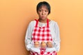 Middle age african american woman wearing baker apron holding muffins puffing cheeks with funny face