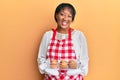 Middle age african american woman wearing baker apron holding muffins winking looking at the camera with sexy expression, cheerful
