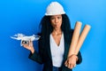 Middle age african american woman wearing safety helmet holding blueprints and drone making fish face with mouth and squinting