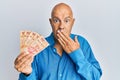 Middle age bald man holding mexican pesos covering mouth with hand, shocked and afraid for mistake