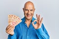 Middle age bald man holding mexican pesos doing ok sign with fingers, smiling friendly gesturing excellent symbol