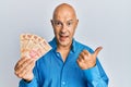 Middle age bald man holding mexican pesos pointing thumb up to the side smiling happy with open mouth