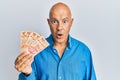 Middle age bald man holding mexican pesos scared and amazed with open mouth for surprise, disbelief face