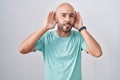 Middle age bald man standing over white background trying to hear both hands on ear gesture, curious for gossip Royalty Free Stock Photo