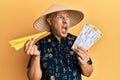 Middle age bald man wearing traditional asian hat holding boarding pass angry and mad screaming frustrated and furious, shouting Royalty Free Stock Photo