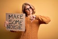 Middle age beautiful blonde activist woman holding banner with make some noise message with angry face, negative sign showing