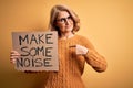 Middle age beautiful blonde activist woman holding banner with make some noise message with surprise face pointing finger to