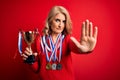 Middle age beautiful blonde successful woman wearing medals holding trophy with open hand doing stop sign with serious and