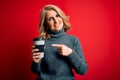 Middle age beautiful blonde woman drinking takeaway cup of coffee over red background very happy pointing with hand and finger Royalty Free Stock Photo