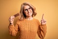 Middle age beautiful blonde woman eating energetic protein bar over yellow background surprised with an idea or question pointing Royalty Free Stock Photo