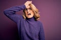 Middle age beautiful blonde woman wearing casual purple turtleneck sweater and glasses surprised with hand on head for mistake, Royalty Free Stock Photo