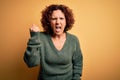 Middle age beautiful curly hair woman wearing casual sweater over isolated yellow background angry and mad raising fist frustrated Royalty Free Stock Photo