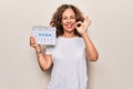 Middle age beautiful woman holding climatology calendar showing cloudy and rainy weather doing ok sign with fingers, smiling