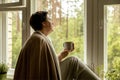 Middle age beautiful woman sitting on windowsill, drinking tea, dreaming. 50-year-old woman relaxing with cup of tea Royalty Free Stock Photo