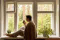 Middle age beautiful woman sitting on windowsill, drinking tea, dreaming. 50-year-old woman relaxing with cup of tea Royalty Free Stock Photo