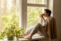 Middle age beautiful woman sitting on windowsill, drinking tea, dreaming. 50-year-old woman relaxing with cup of tea Royalty Free Stock Photo