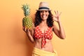 Middle age beautiful woman wearing bikini and summer hat holding pineappel doing ok sign with fingers, smiling friendly gesturing Royalty Free Stock Photo