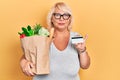 Middle age blonde woman holding groceries and credit card relaxed with serious expression on face Royalty Free Stock Photo