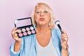 Middle age blonde woman holding makeup brush and blush puffing cheeks with funny face