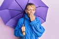 Middle age blonde woman holding purple umbrella covering mouth with hand, shocked and afraid for mistake