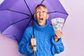 Middle age blonde woman holding umbrella and russian ruble banknotes angry and mad screaming frustrated and furious, shouting with Royalty Free Stock Photo