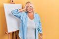 Middle age blonde woman standing by painter easel stand surprised with hand on head for mistake, remember error Royalty Free Stock Photo